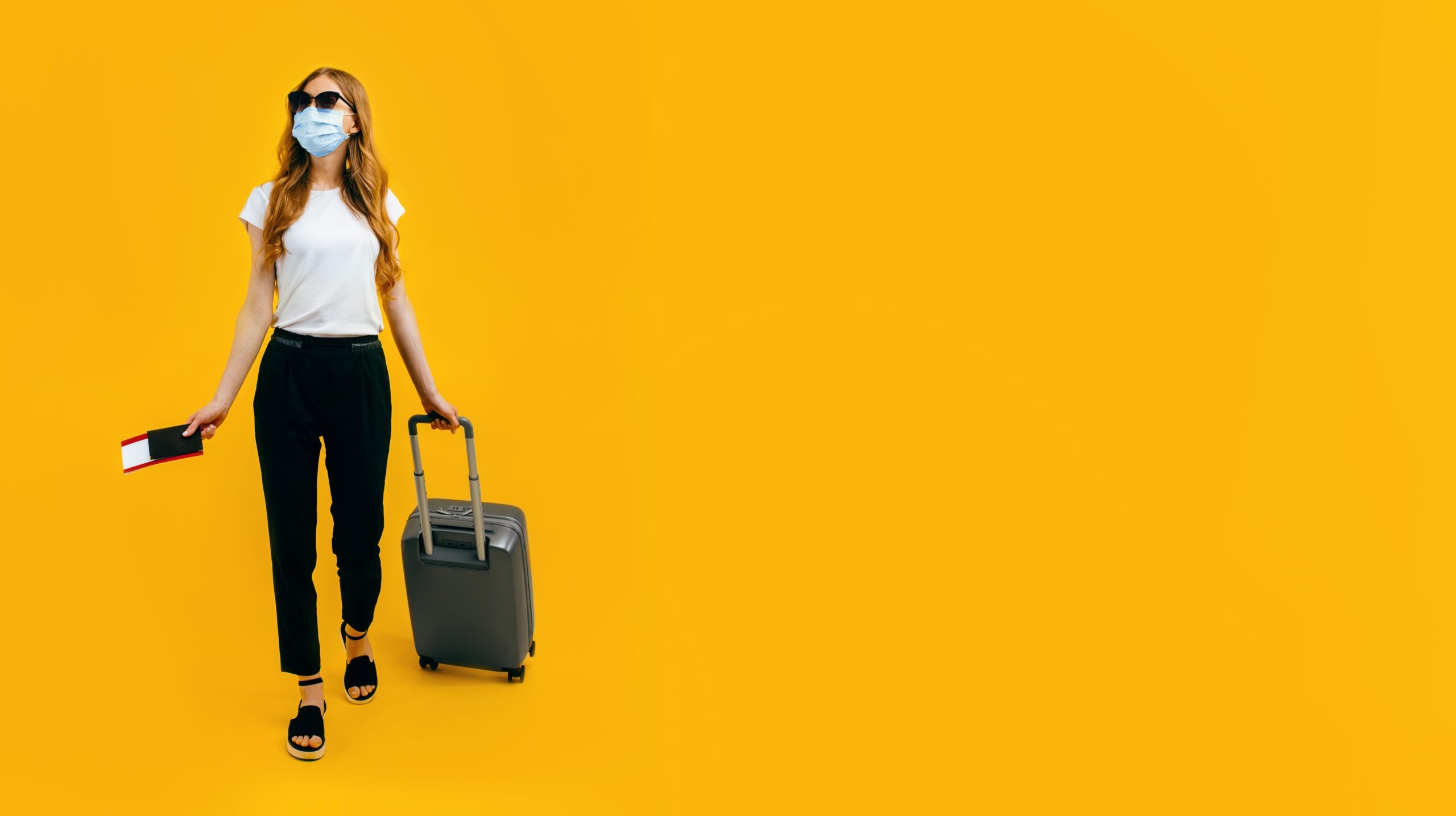 A female passenger in a medical protective mask on her face, with a suitcase and passport, goes abroad. Concept of travel, quarantine and coronavirus